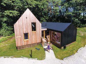 A Burnbake forest lodge. One part of the lodge is made of natural coloured wood with two glass windows and the other half of black wood with a floor length glass window.