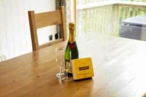 A champagne bottle. empty champagne glass and small yellow box of chocolates sit on a wooden table.