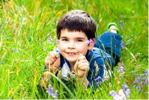 A boy with brown hair lays on his stomach in the grass, smiling at the camera and holding two delicate purple flowers.