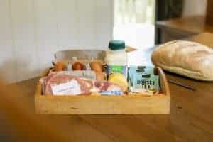 A wooden box containing a pack of bacon, a small bottle of milk, teabags and eggs sits on a table next to a load of bread and a knife.