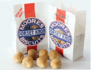 Two white paper packets are decorated with a vertical red stripe and a blue circle which displays the phrase "Moores Dorset Knob Biscuits". Seven small biscuits resembling bread rolls sit in front of the packets.