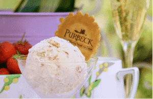 A glass bowl contains a round scoop of ice cream and a wafer embossed with the Purbeck Icecream logo, depicting the silhouette of Corfe Castle. In the background is a glass of champagne and a bowl of strawberries, plus a purple bucket of flowers.