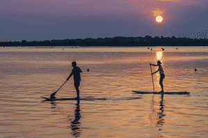 A man and woman paddle on still water, on paddle boards, under a blue and orange sunset, with birds in the background.