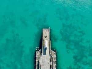 Aerial view of a grey stone pier, sitting on a turquoise sea.