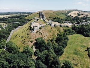 An aerial view of Corfe Castle. A ruined castle made of grey stone sits on top of a grassy hill, surrounded by trees. 