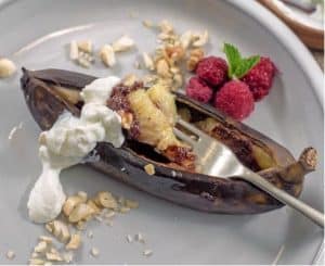 A brown banana skin is sliced lengthways and a silver fork holds banana, chocolate and cream. A grey plate containing four raspberries holds the banana.
