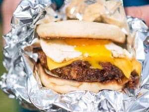 A sausage patty, topped with a fried egg and melted cheese slice, inside an english muffin roll. The egg yolk drips from the egg, surrounded by a tin foil wrapping.