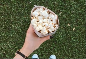 A person wearing a black watch strap holds a brown paper bag full of popcorn, against a background of green grass.