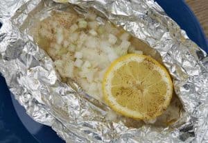 White, baked fish and a slice of lemon inside a silver tin foil wrapping which has been opened to reveal the fish.