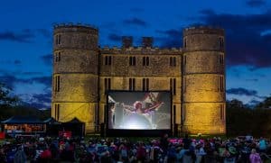 Lulworth Castle at night. A castle with two round towers displays a cinema screen and a crowd of people gathers to watch.