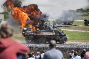 A dark green military armoured tank crawls along a grass track. An explosion of orange flames and black smoke is seen behind the tank, as crowds look on.