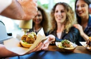 A woman with curly blonde hair holds out her hand, preparing to receive a bun containing three falafel balls, onto which a man is squeezing sauce.
