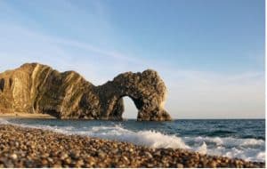 Durdle Door on a sunny day. An archway of rock surrounded by dark blue sea. Waves crash on a pebble beach.