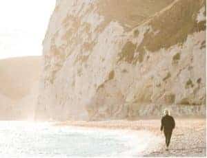 A blonde haired woman walks along the shore of a beach, under a white cliff, on a bright day.