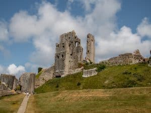 The ruins of Corfe Castle on a sunny day. The castle sits on top of a grassy hill, with a grey path leading up to it.