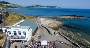 An aerial view of the Marine Theatre, Lyme Regis. A white stone building with a grey roof, next to the sea.