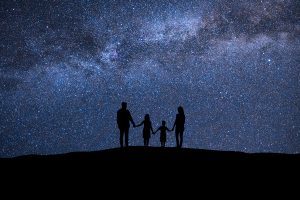 The silhouette of a family of two adults and two children hold hands in front of a blue starry sky.