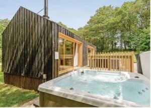 A hot tub with clear blue water bubbles in front of a Burnbake lodge with dark wood panel walls, surrounded by green oak trees.