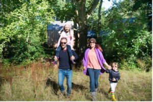 A father walks with his daughter on his shoulders and a mother holds her son's hand, in a field on a sunny day.