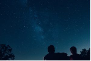 The silhouette of a couple with their backs to the camera, against a dark blue starry sky at night. The milky way is faintly visible.