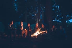 Six adults face the camera, behind a glowing bonfire, in a dark forest.