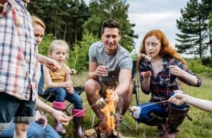 A family of 6 toasts marshmallows around a small bonfire in a green field, surrounded by pine trees.