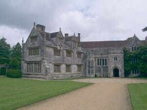 A Tudor manor house with a light grey stone exterior, surrounded by a gravel pathway and green grass.