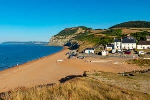 A view of the Golden Cap cliffs above a sandy beach and white pub buildings, under a bright blue sky.