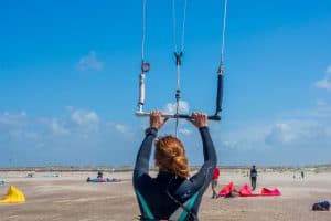 A woman with light brown hair holds the handle of a kitesurfing kite on a beach, under a blue sky.