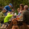 Family enjoy picnic and barbecue food around a brown picnic bench.